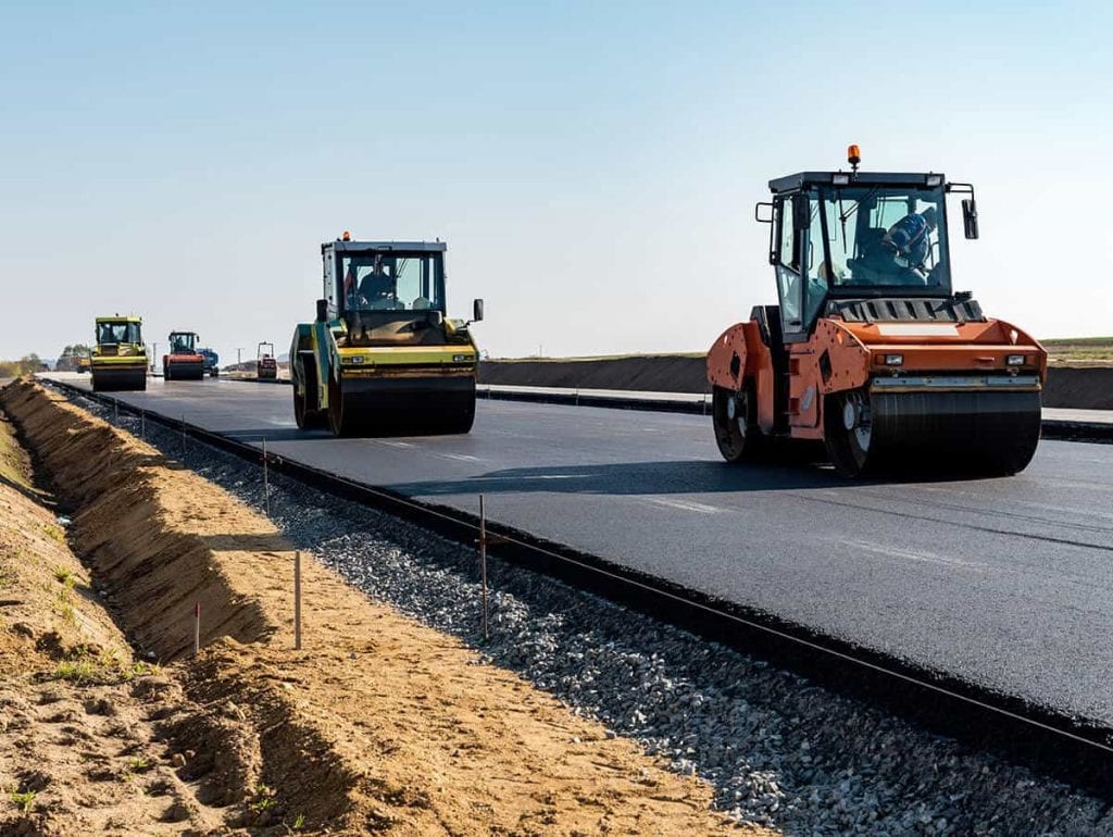 Several road rollers and construction vehicles are paving and smoothing fresh commercial asphalt on a wide road under clear skies, with sand and gravel along the edges—perfect for residential asphalt projects in Wilson County.
