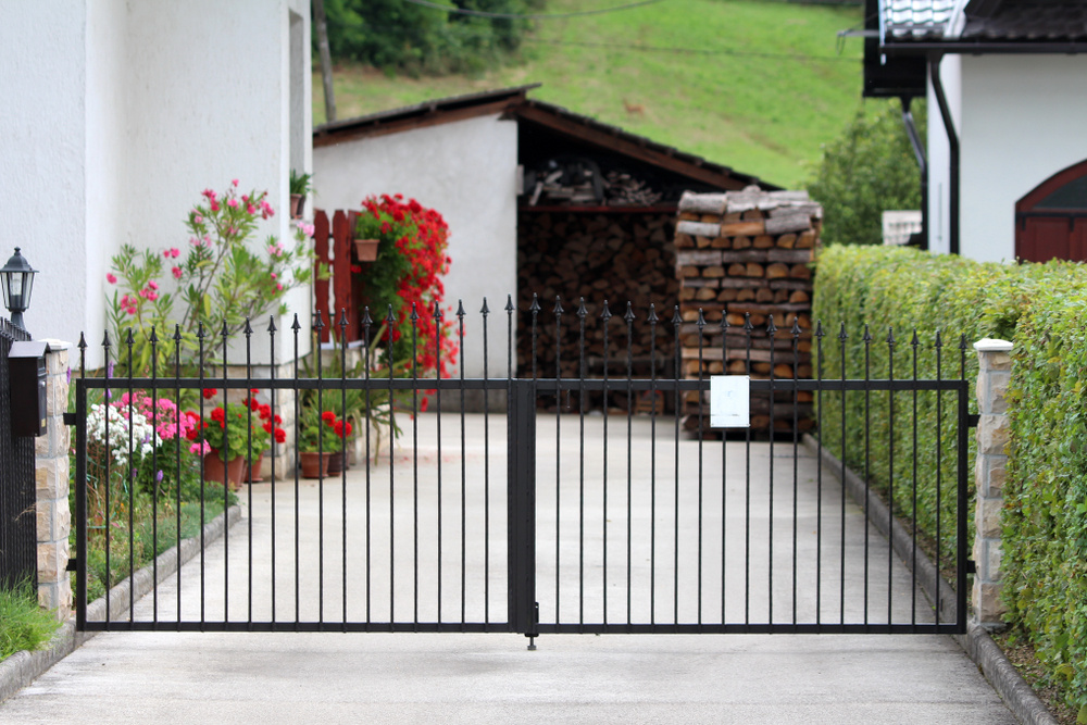 A black metal gate blocks a driveway with residential asphalt, leading to a shed stacked with firewood. Red and pink flowers line the left side, and green hedges border the path. White buildings are visible on both sides.