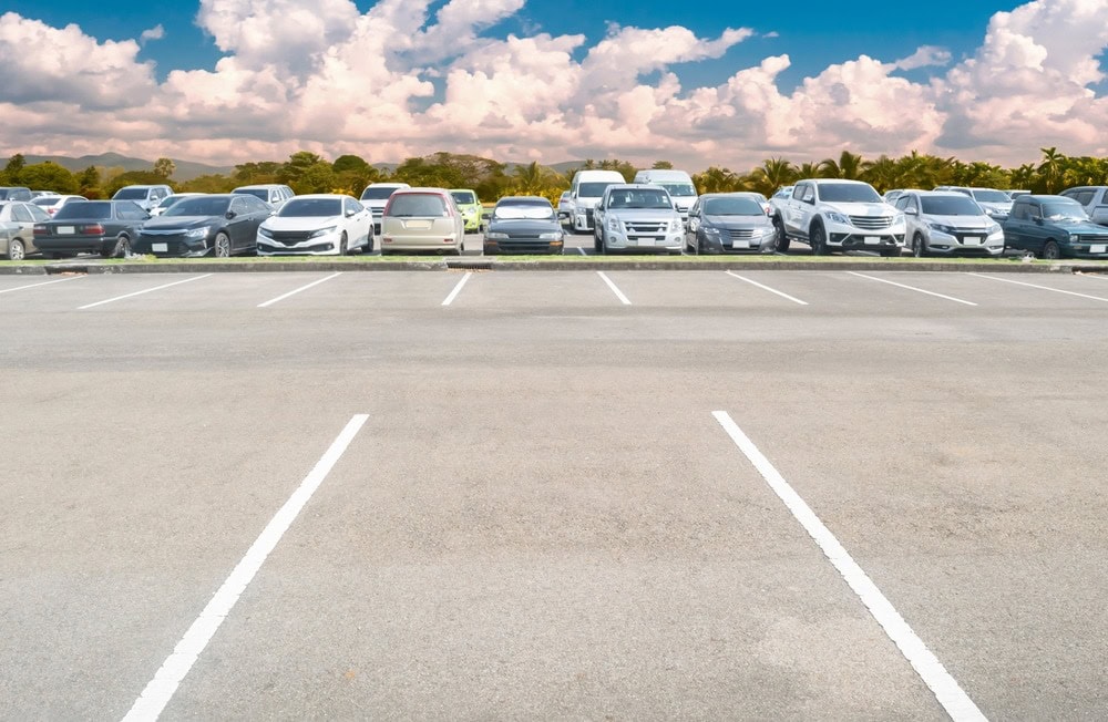 A row of parked cars fills the spaces at the edge of a parking lot, while several parking spots in the foreground are empty—freshly finished with sealcoating Wilson County. Trees and a cloudy sky are visible in the background.
