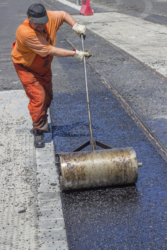 A worker in orange overalls and a cap uses a heavy metal roller to smooth fresh driveway asphalt on a street, near tram tracks and concrete pavement.