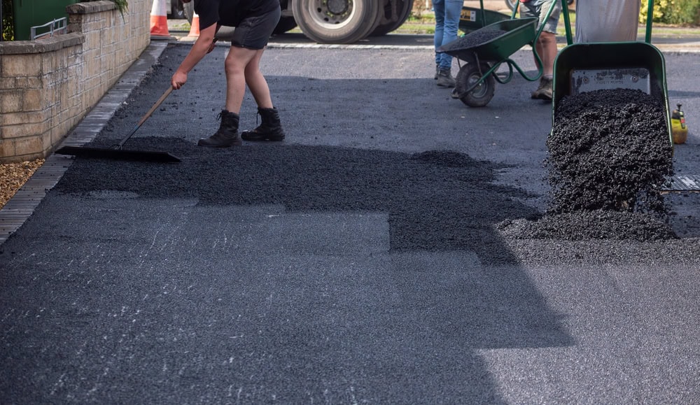 Two workers are laying fresh asphalt on a driveway. One uses a rake to spread the asphalt, while the other stands nearby with a wheelbarrow full of more material. The fresh asphalt is visibly darker than the older section.