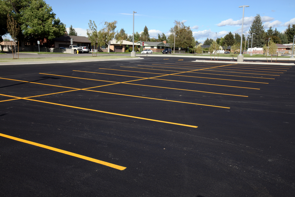 An empty parking lot with freshly painted yellow lines under a clear blue sky, surrounded by trees and buildings in the background.