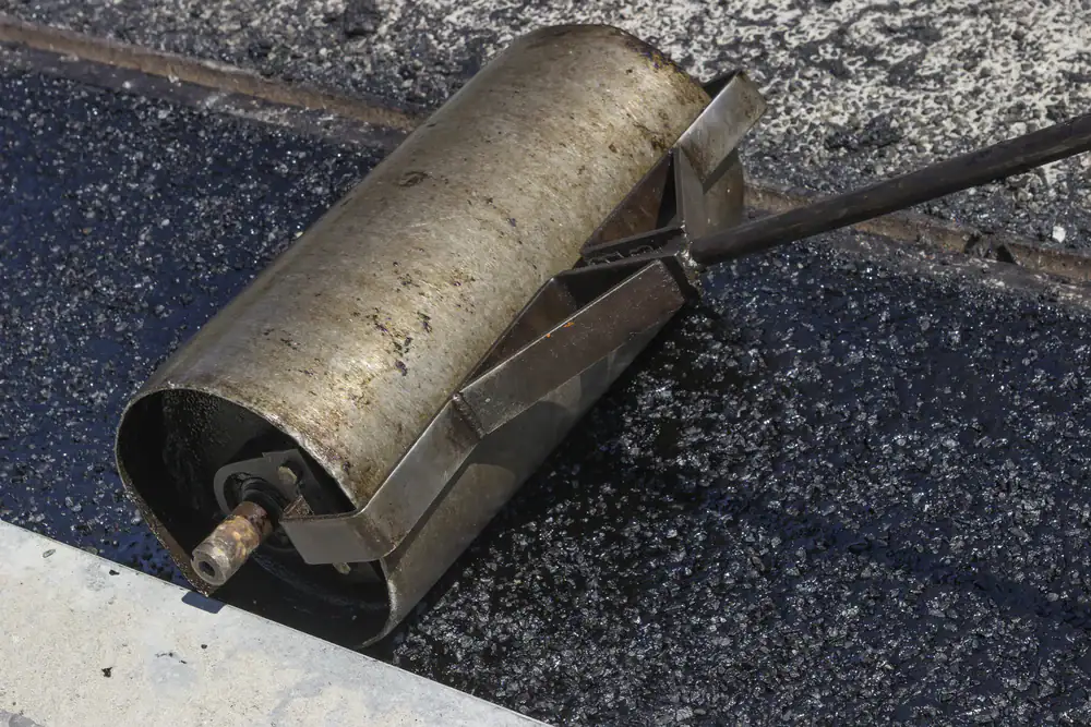 A close-up of a metal roller being used to smooth hot asphalt on a road, with fresh black asphalt and concrete curb visible.