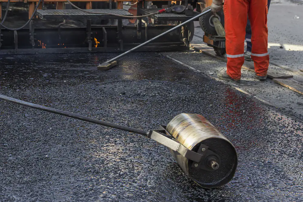 A construction worker in orange overalls smooths fresh asphalt with a heavy metal roller on a road under repair. The surface is shiny and dark, and machinery and tools are visible in the background.