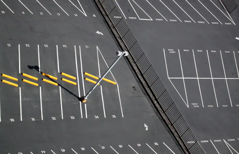 A nearly empty parking lot viewed from above, with white-lined parking spaces, yellow bumpers, a single streetlight casting a shadow, and a fence running diagonally across the lot.