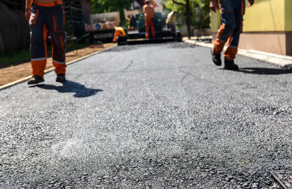 Workers in orange uniforms are paving a road with fresh black asphalt on a sunny day. The focus is on the new asphalt surface, with workers and paving equipment visible in the background.