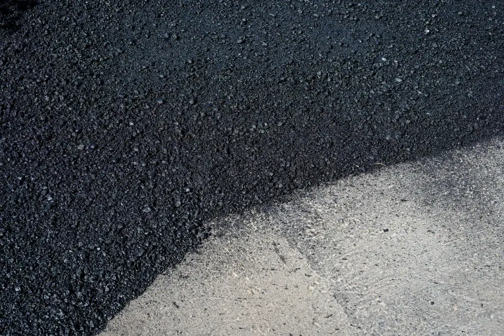 A close-up view showing fresh black asphalt partially covering an older, light gray concrete surface, illustrating a road or pavement resurfacing process.