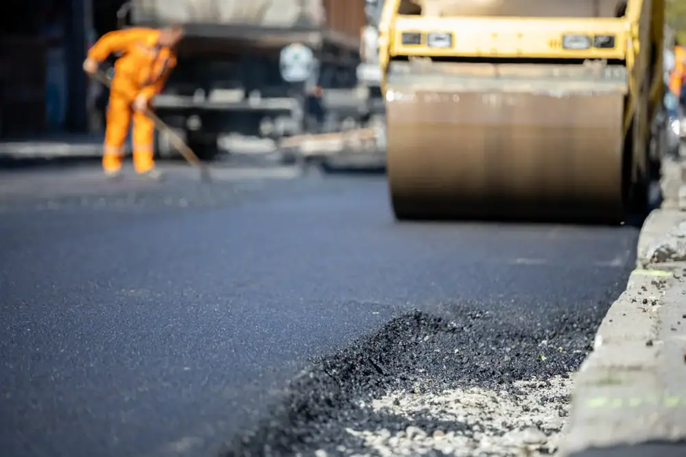 A road roller smooths freshly laid asphalt on a street, while a worker in an orange uniform uses a tool in the background. The scene shows ongoing road construction.