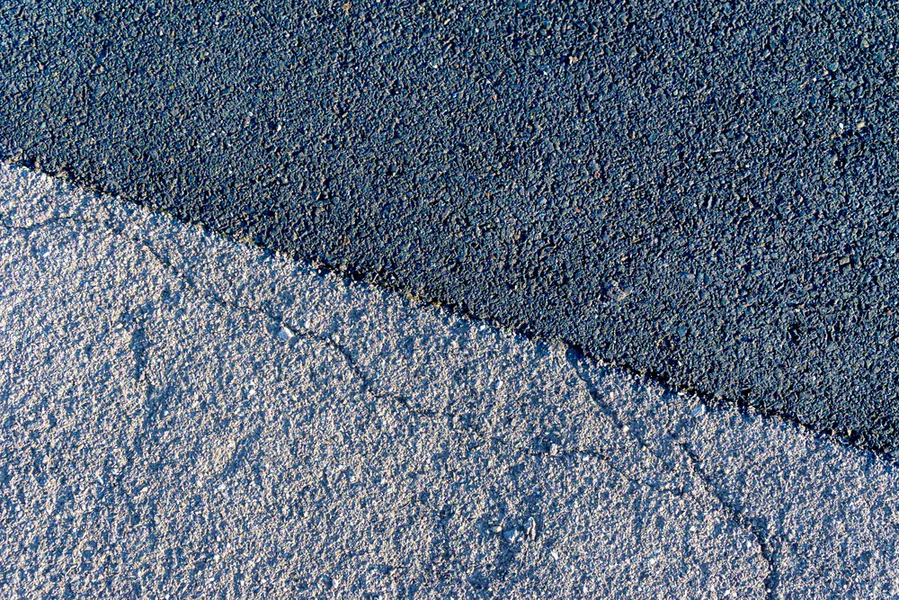 A close-up view of two contrasting pavement surfaces: a smooth, dark asphalt on the top right, and a rough, light gray concrete on the bottom left, separated by a diagonal line.
