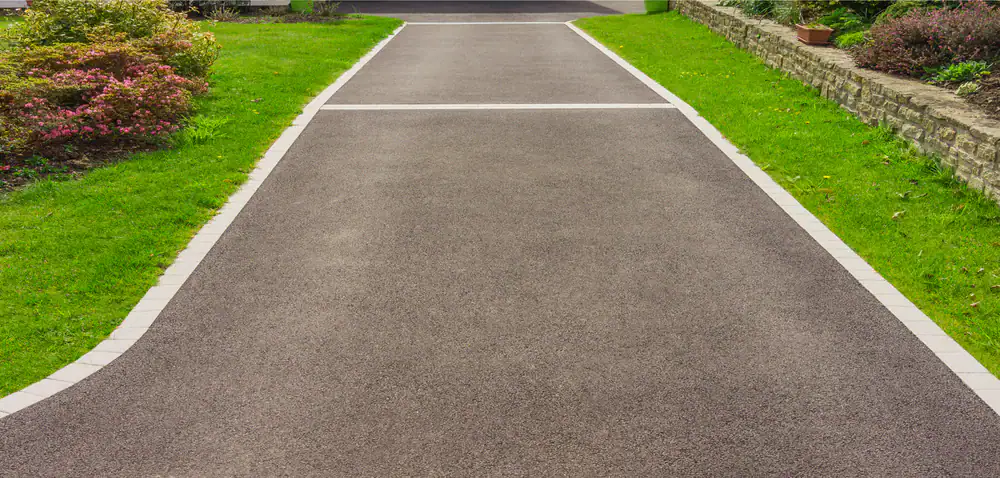 A smooth, paved driveway bordered with white bricks runs between green grass and garden beds with bushes and a stone wall on one side.