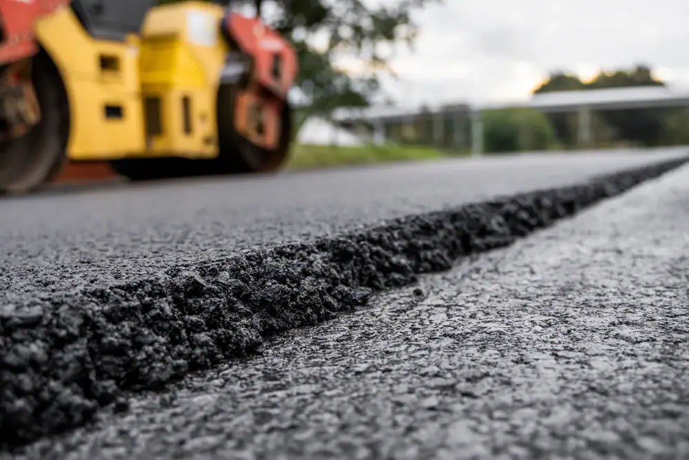 Close-up of freshly laid asphalt on a road, with a blurred yellow and red steamroller in the background and trees visible along the roadside.